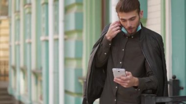 Slow Motion genç adam yeşil binanın yanında duruyor, koruyucu maskeyi çıkarıyor ve akıllı telefonuna bakıyor.
