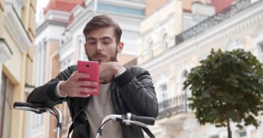 Sırt çantalı bisikletli kurye akıllı telefonundaki fotoğraflara bakıyor. Bisikletli genç adam sokakta duruyor, akıllı telefon kullanıyor ve gülümsüyor. Sinema 4K 60fps video