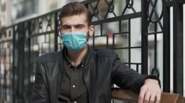 Tıbbi koruyucu maskeli genç adam bankta oturuyor ve sana bakıyor. Covid konsept sağlık ve güvenlik, N1H1 koronavirüs karantinası, bir sonraki dalga covid virüs koruması
