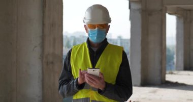 Kasklı ve tıbbi koruyucu maskeli işçiler inşaat alanında duruyor ve akıllı telefonuna veri girişi yapıyor. Sinema 4K 60fps video