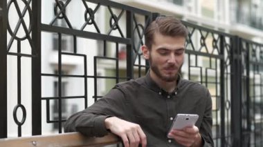 Genç adam bankta oturur ve akıllı telefonundaki fotoğraflara bakar, gülümser ve keyfi yerindedir.
