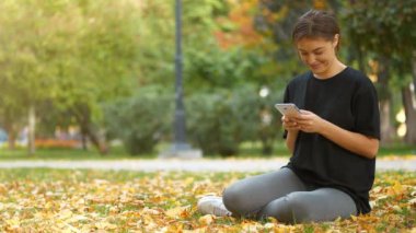 Genç ve güzel bir kız sonbahar parkında çimlerde oturur, akıllı telefonundaki fotoğraflara bakar ve bakar, gülümser, güler ve keyfi yerindedir.