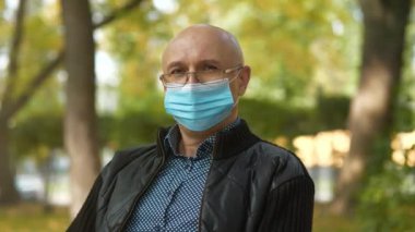 Mavi tıbbi koruyucu maskeli, gözlüklü kel bir adam bankta oturur ve kameraya bakar. Arka planda arabalar yolda gidiyor.