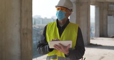 Beyaz başlıklı ve tıbbi koruyucu maskeli inşaatçı inşaat alanında duruyor ve tablet bilgisayarına veri giriyor. Sinema 4K 60fps video