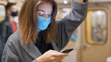 Tıbbi maskeli bir kadın, akıllı telefonlu bir kadın metro vagonunda duruyor. Koruyucu maskeli diğer yolcular metro vagonuna biniyor.