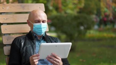 Mavi tıbbi koruyucu maskeli kel adam bankta oturuyor ve tablet bilgisayarla iletişim kuruyor.