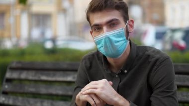 Mavi tıbbi koruyucu maskeli genç bir adam bankta oturur ve kameraya bakar.