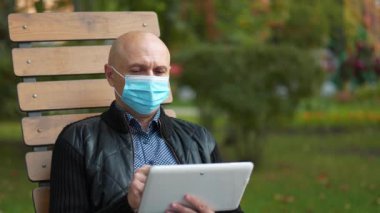 Mavi koruyucu maskeli kel adam bankta oturur ve tablet bilgisayar kullanır.