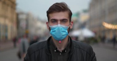 Mavi tıbbi koruyucu maskeli genç adam sokakta dikiliyor ve kameraya bakıyor, kırmızı lazer ışını alnında yükseliyor ve parlıyor, adam gözlerini kapatıyor, Cinema 4K 60fps video