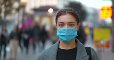 Kalabalık bir sokakta mavi tıbbi koruyucu maskeli genç bir kadın kameraya bakıyor. Sinema 4K 60fps panoramik video