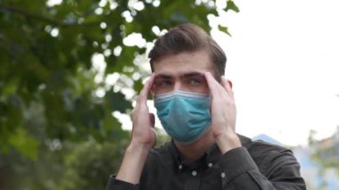 Tıbbi koruyucu maskeli genç adam kafasına masaj yapıyor. Kendini iyi hissetmiyor ve başı ağrıyor.