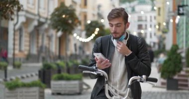 Tıbbi koruyucu maskeli genç adam sokakta bisikletle dikiliyor ve akıllı telefondan konuşuyor. Sırt çantalı bisikletli kurye akıllı telefonla iletişim kuruyor. Sinema 4K 60fps video