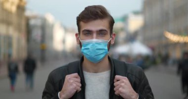 Mavi tıbbi koruyucu maskeli genç adam sokakta dikiliyor ve kameraya bakıyor. Sinema 4K 60fps video