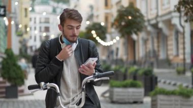 Bisiklet çantalı bisikletli kurye sokakta duruyor ve akıllı telefondan konuşuyor. Genç adam tıbbi koruyucu maskeni çıkar ve akıllı telefondan konuşmaya başla.