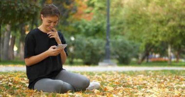 Genç ve güzel bir kız sonbahar parkında çimlerde oturur, akıllı telefon kullanır, fotoğraflara bakar ve bakar, gülümser ve güler. Elektrikli scooterlı iki adam yol boyunca araba kullanıyor. Sinema 4K 60fps video