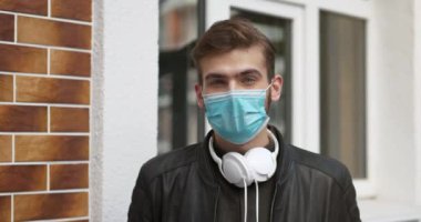 Tıbbi koruyucu maskeli, kulaklıklı genç adam evin penceresinin yanında durup kameraya bakıyor.