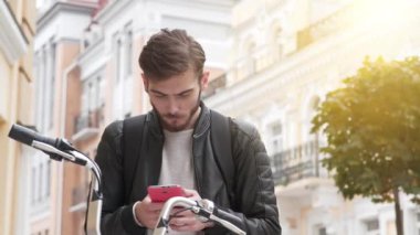 Sırt çantalı bisikletli kurye akıllı telefonuna bakar ve zaferi kutlar ve şiddet dolu duygular gösterir. Genç adam bisikletle sokakta duruyor ve akıllı telefon kullanıyor.
