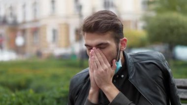 Koruyucu maskeli hasta genç adam şehir meydanında bir bankta oturuyor, öksürüyor ve burnunu siliyor.