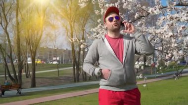 Güneş gözlüklü adam parkta akıllı telefondan konuşuyor. Akıllı telefonlu kırmızı beysbol şapkalı adam.