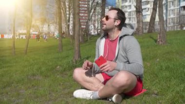 Güneş gözlüklü yetişkin adam şehir parkındaki çimenlerde oturur ve müzik dinler, arka planda güzel bir güneş vardır.