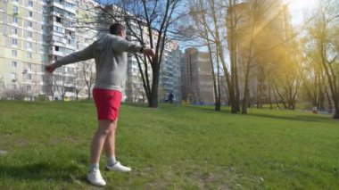 Adam parkta çimenler üzerinde fiziksel egzersiz yapıyor, arka planda güzel bir güneş.