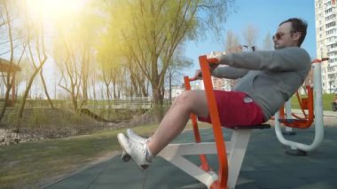 Adam spor sahasında fiziksel egzersiz yapıyor. Sporcu yorucu bir egzersiz yapıyor. Açık hava spor salonunda gözlüklü aktif bir adam, arka planda güzel bir güneş.