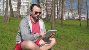 Adam şehir parkında tablet bilgisayar kullanıyor. Adam çimlerin üzerine oturur ve tablet bilgisayarına bakar, sayfaları çevirir, makale okur.