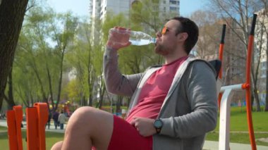 Sporcu oturur ve şişeden su içer. Gözlüklü adam spor salonunda su içiyor.