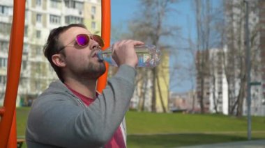 Gözlüklü adam dışarıda spor salonunda su içiyor. Sporcu oturur ve şişeden su içer.