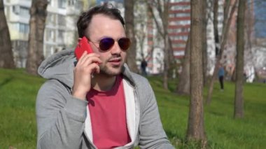 Gözlüklü kızgın adam akıllı telefondan konuşuyor. Kırmızı akıllı telefonlu duygusal bir adam parktaki çimlerde oturur, konuşur ve el kol hareketi yapar.