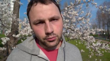 Adam kameraya bakar ve konuşur. Yetişkin bir adam şehir parkındaki kayısı ağacının yanında duruyor ve kameraya doğru konuşuyor.