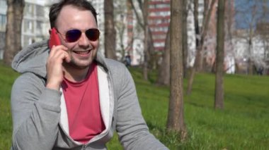 Adam akıllı telefondan konuşuyor. Şehir parkında kırmızı akıllı telefonu olan adam konuşuyor ve el kol hareketi yapıyor.
