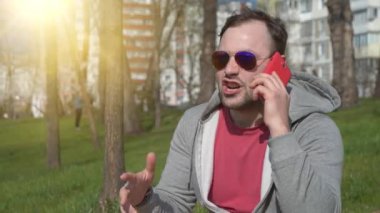 Kırmızı akıllı telefonlu duygusal bir adam parktaki çimlerin üzerinde oturur, konuşur ve el kol hareketi yapar. Gözlüklü kızgın adam akıllı telefonda konuşur, arka planda güzel bir güneş.