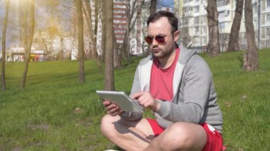 Adam şehir parkında tablet bilgisayar kullanıyor. Adam çimlerin üzerinde oturur ve tablet bilgisayarına bakar. Arka planda güzel bir güneş vardır.