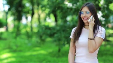 Gözlüklü güzel kadın cep telefonuyla konuşuyor. Çekici kadın halka açık bir bahçede duruyor ve cep telefonuyla konuşuyor. Beyaz akıllı telefonlu bir kadın.