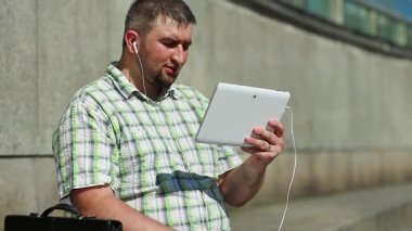 İşadamı tablet bilgisayarla iletişim kuruyor. Tabletli adam video kaset aracılığıyla iletişim kuruyor. Evrak çantalı, kareli gömlekli şişman bir adam elinde tabletle kulaklık tutuyor.