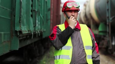 Trenlerin yanında taşınabilir radyo vericisi olan bir demiryolu işçisi. Demiryolu çalışanı, nakliye istasyonunun yanındaki demiryolu hattında portatif radyo vericisi aracılığıyla konuşuyor. İşçi radyo istasyonu aracılığıyla iletişim kuruyor.