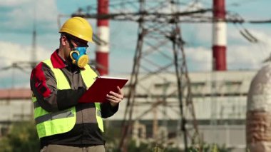 Elektrik santralindeki gaz maskeli adam iş belgelerine not alıyor. Elektrik mühendisliği uzmanı. Solunum cihazında ve sarı kaskette. Belgelerde yazıyor.