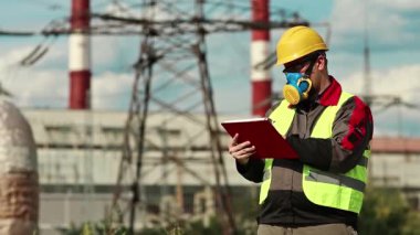 Elektrik santralindeki gaz maskeli adam iş belgelerine not alıyor. Elektrik mühendisliği uzmanı. Solunum cihazında ve sarı kaskette. Belgelerde yazıyor.