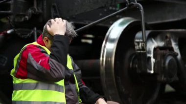Demiryolu raylarında stresli demiryolu işçisi demiryolu hattında büyük metal tekerleğin yanında oturuyor. Kırmızı şapkalı yorgun demiryolu çalışkan bir günün ardından yük vagonunun yanında oturur.