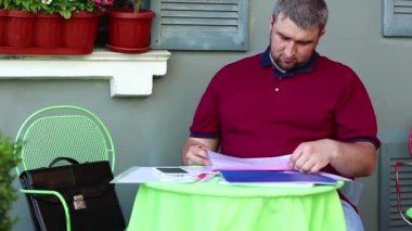 Sinirli ve sinirli iş adamı restoranda masada oturuyor ve belgelerini karşılaştırıyor. Stres altındaki kızgın adam yaz kafesinde oturur ve iş belgeleriyle çalışır.