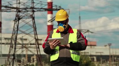 Elektrik santralindeki tablet bilgisayarlı gaz maskeli adam. Güç mühendisliği uzmanı solunum cihazında ve sarı kasketli ve kişisel bilgisayarlı ısı santralinde çalışıyor.