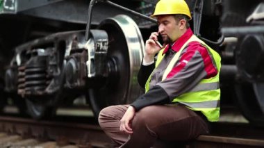 Demiryolu işçisi cep telefonuyla konuşuyor. Demiryolu çalışanı iş kıyafetleri içinde yük vagonunun yanında oturur ve cep telefonuyla konuşur.