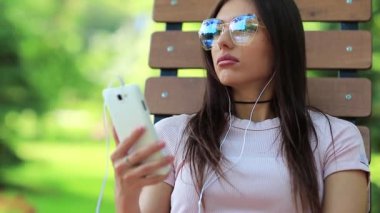 Çekici kız şehir parkında bankta oturur ve müzik dinler. Genç ve güzel bir kadın halka açık bir bahçede bankta oturur ve akıllı telefondan müzik dinler.
