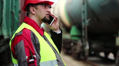 Demiryolu işçisi tren yakınlarında cep telefonuyla konuşuyor. Demiryolu çalışanı demiryolu hattında çalışıyor. Cep telefonuyla konuşuyor. İşyerinin müdürü, nakliye istasyonundaki akıllı telefon aracılığıyla iletişim kuruyor.