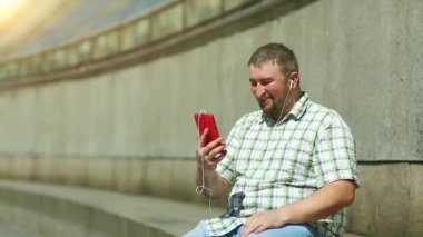 Komik adam akıllı telefondan konuşuyor. Akıllı telefonlu neşeli adam Skype üzerinden iletişim kuruyor ve el kol hareketi yapıyor. Elinde kırmızı cep telefonu olan, kareli tişörtlü, şişman, şen şakrak adam.