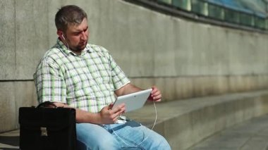 İşadamı tablet bilgisayarla iletişim kuruyor. Tabletli adam video kaset aracılığıyla iletişim kuruyor. Evrak çantalı, kareli gömlekli şişman bir adam elinde tabletle kulaklık tutuyor.