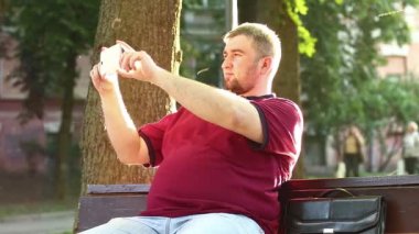 İş adamı bankta oturur ve akıllı telefondaki videoları kaydeder. Şişman adam cep telefonuyla fotoğraf çekiyor.