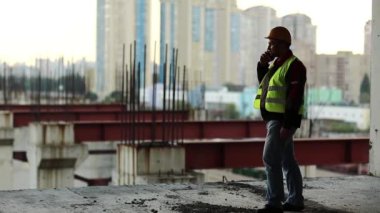 Foreman inşaat alanında cep telefonuyla konuşuyor. Yeşil yelek giyen inşaatçı proje sitesinde telefonla konuşuyor