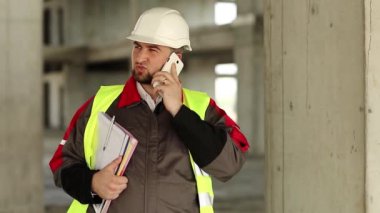 İnşaat alanında akıllı telefonu olan bir inşaatçı. İnşaatçı, cep telefonuyla proje alanında konuşuyor. İnşaat alanındaki akıllı telefon konuşmalarında sarı başlıklı çalışma belgelerine sahip ustabaşı.